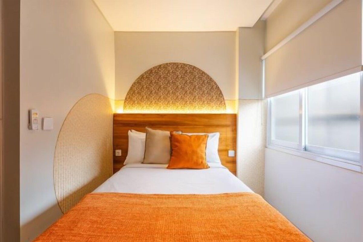 Una habitación de hotel con una cama, un ventanal grande y una lámpara fijada a la pared.