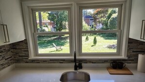 A kitchen with a double sink, white countertops, a window with a view of a backyard, and a brick backsplash.