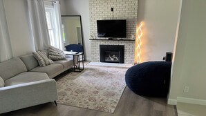 A living room with a grey sofa, a black TV mounted on a brick wall, a fireplace, and a dark bean bag.