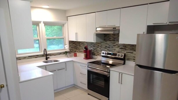 A modern kitchen with white cabinets, a stainless steel refrigerator, a built-in oven, and a window with a view of greenery.