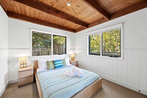 A bedroom with a wooden ceiling, a bed with a patterned bedspread, two bedside tables with lamps, and windows with views of greenery.