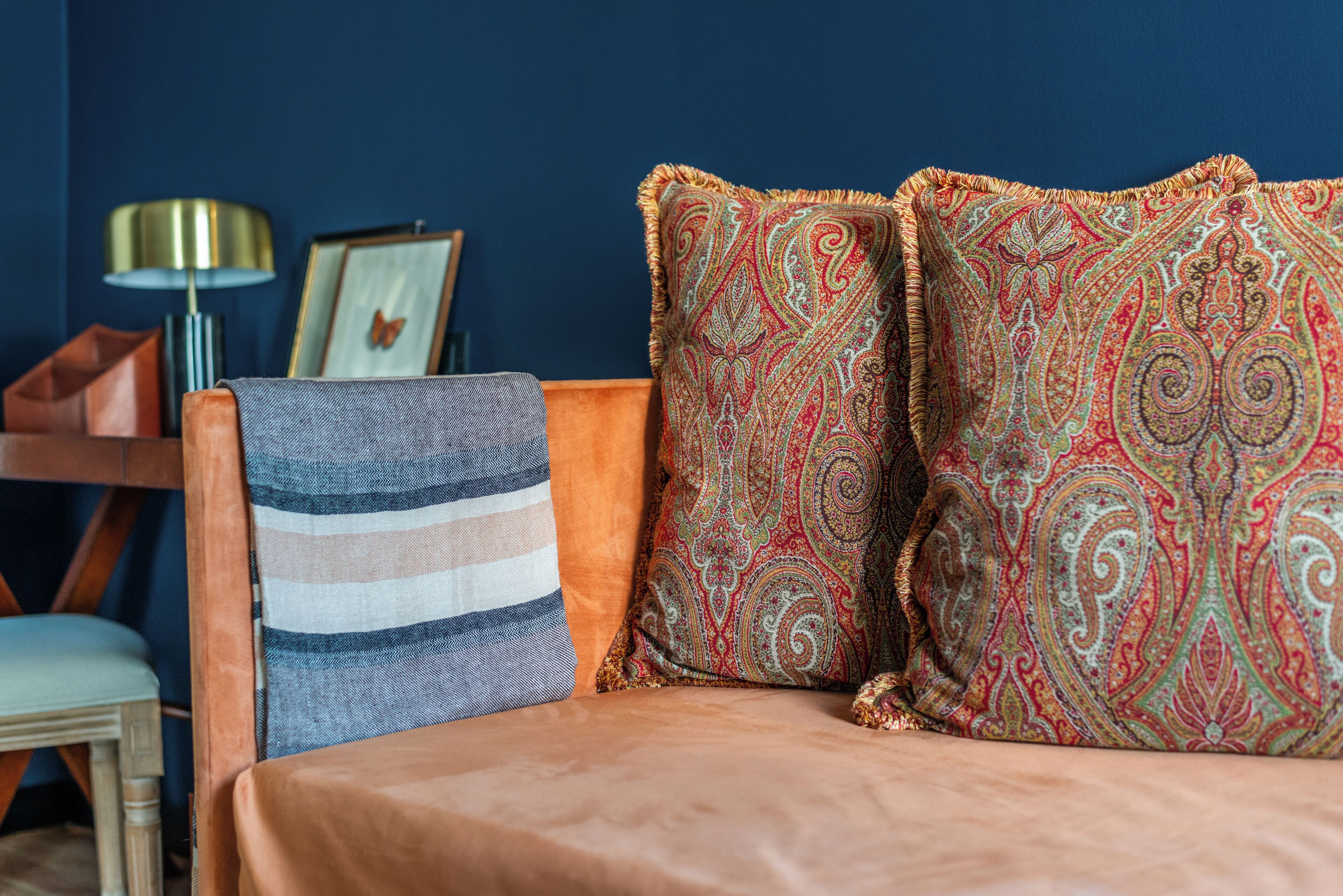 Un canapé agrémenté de coussins à motifs et d’un plaid rayé, une lampe et un tableau encadré sur un mur bleu.