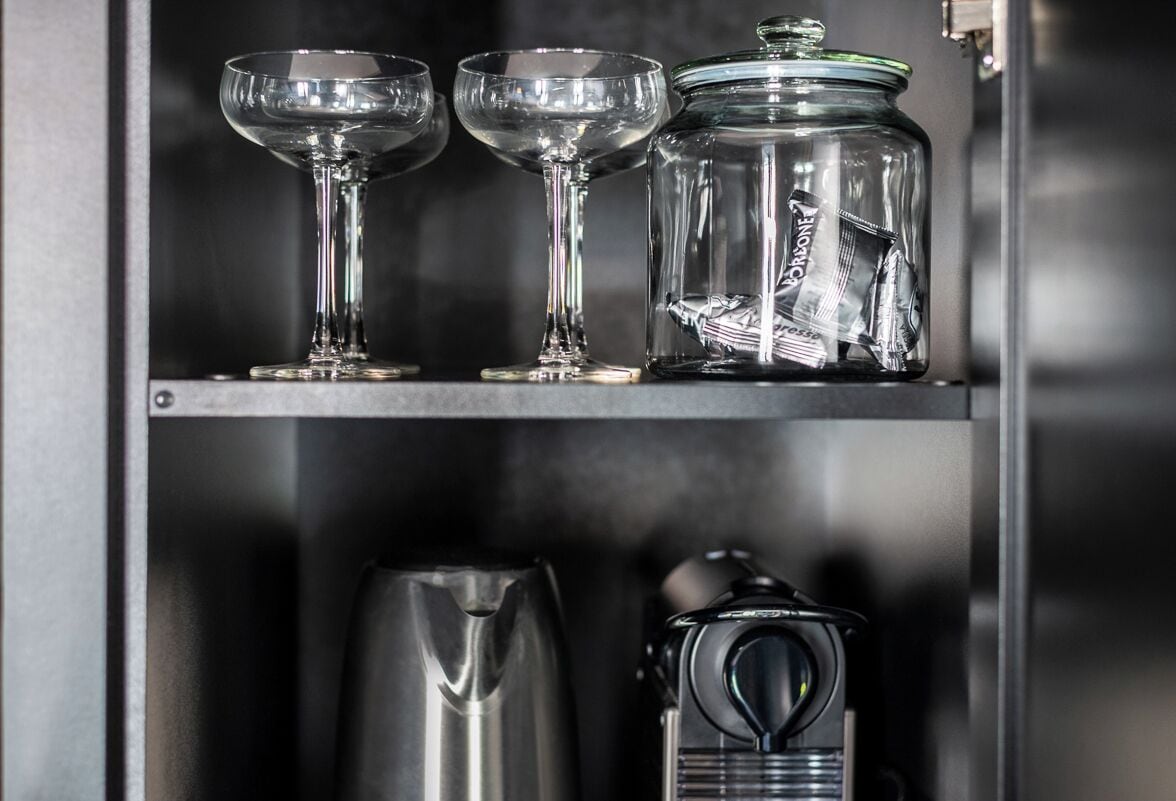 Una estantería con dos copas de vidrio transparentes, un tarro de vidrio con tapa y una cafetera.