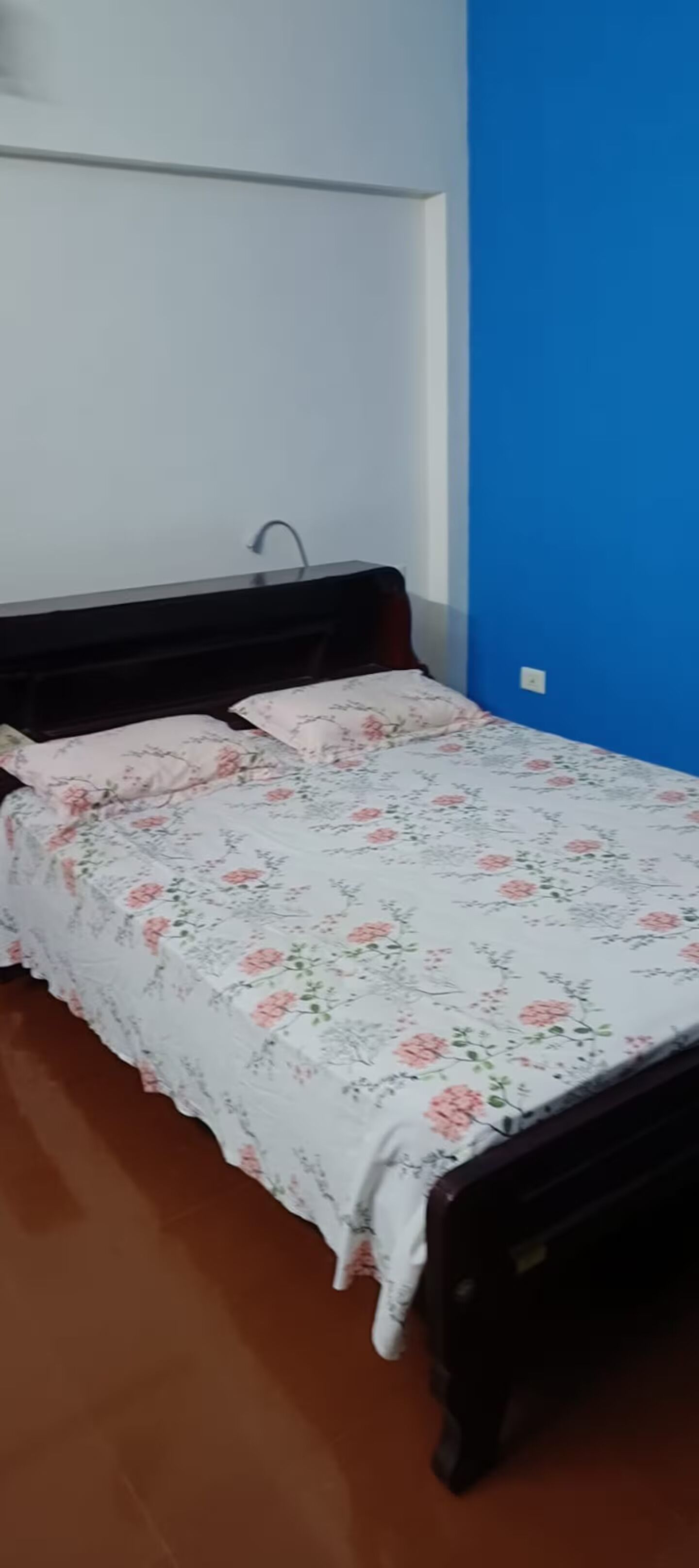 A bedroom with a bed covered in a floral bedspread, a wooden headboard, and a blue wall.