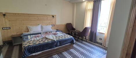 A hotel room with a wooden headboard, a bed with a patterned bedspread, a small table with a chair, and a view of the outdoors through a window with curtains.