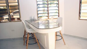 Un coin repas avec une table à plateau en verre, des chaises blanches et une fenêtre équipée de stores horizontaux.