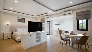A modern hotel room with a bed, TV, white cabinets, a dining table with chairs, and a window.