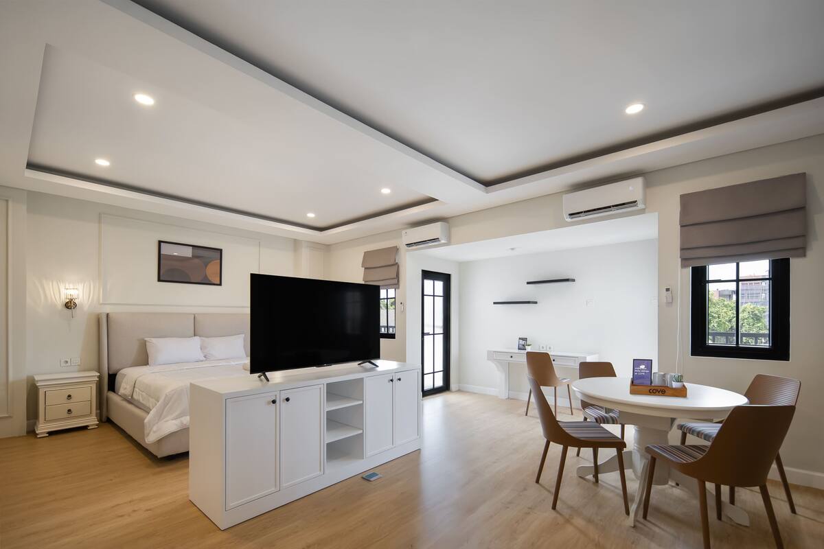 Une chambre d’hôtel moderne équipée d’un lit, d’une télévision, d’armoires blanches, d’une table à manger avec des chaises et d’une fenêtre.