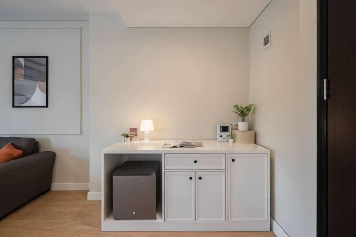 Une pièce moderne avec un canapé gris, un bureau blanc avec une lampe, une petite plante et une porte sombre.