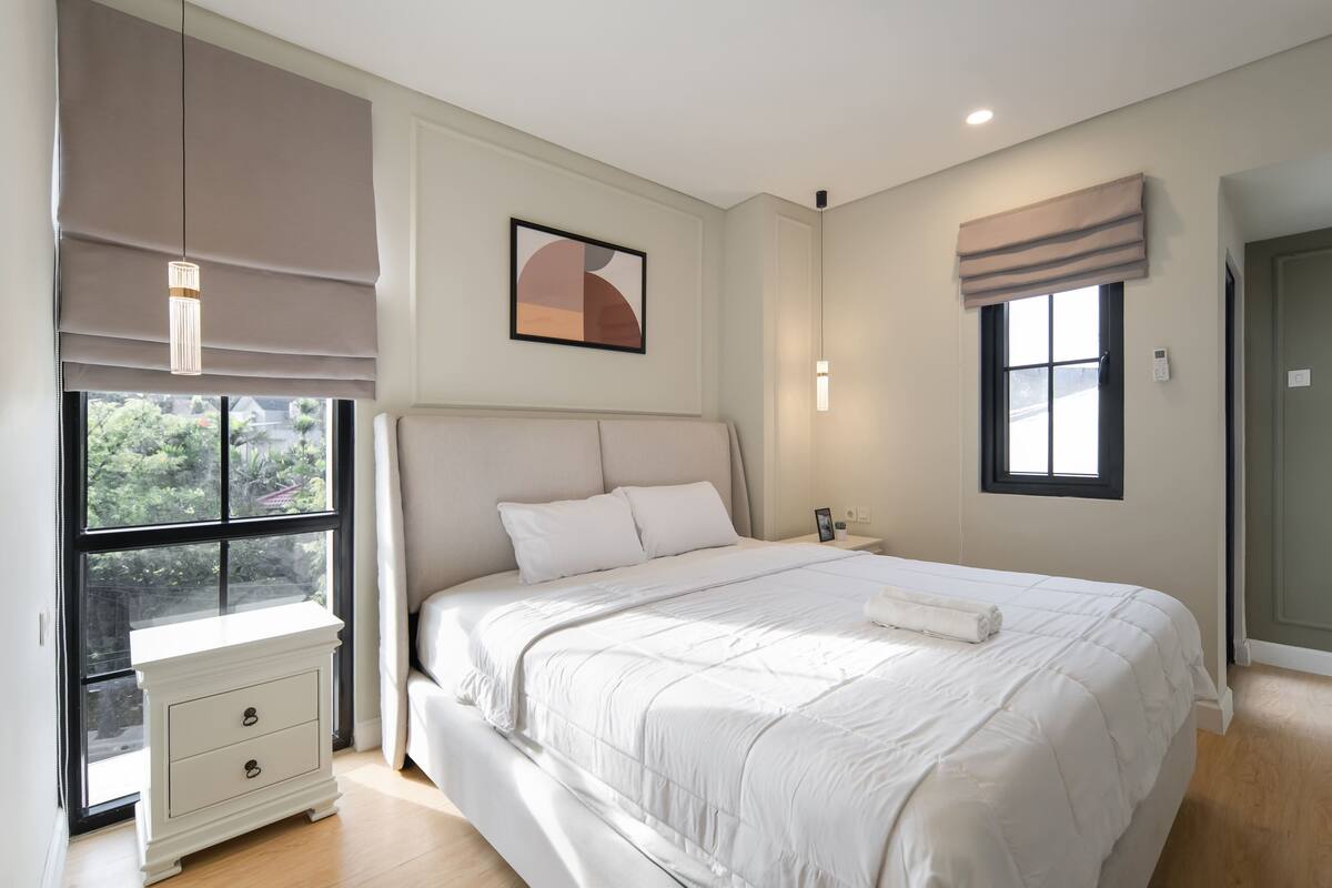 Une chambre avec un grand lit, une table de chevet, une commode, une fenêtre avec des stores et un luminaire fixé au mur.
