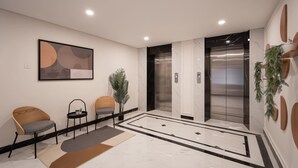 A modern lobby with a marble floor, two wooden chairs, a small table with a plant, and two elevators.