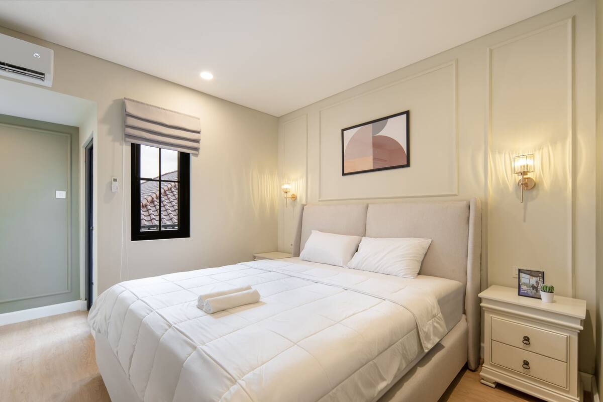 Une chambre à coucher bien rangée, avec un grand lit, des tables de chevet, un tableau accroché au mur et une fenêtre avec des stores.