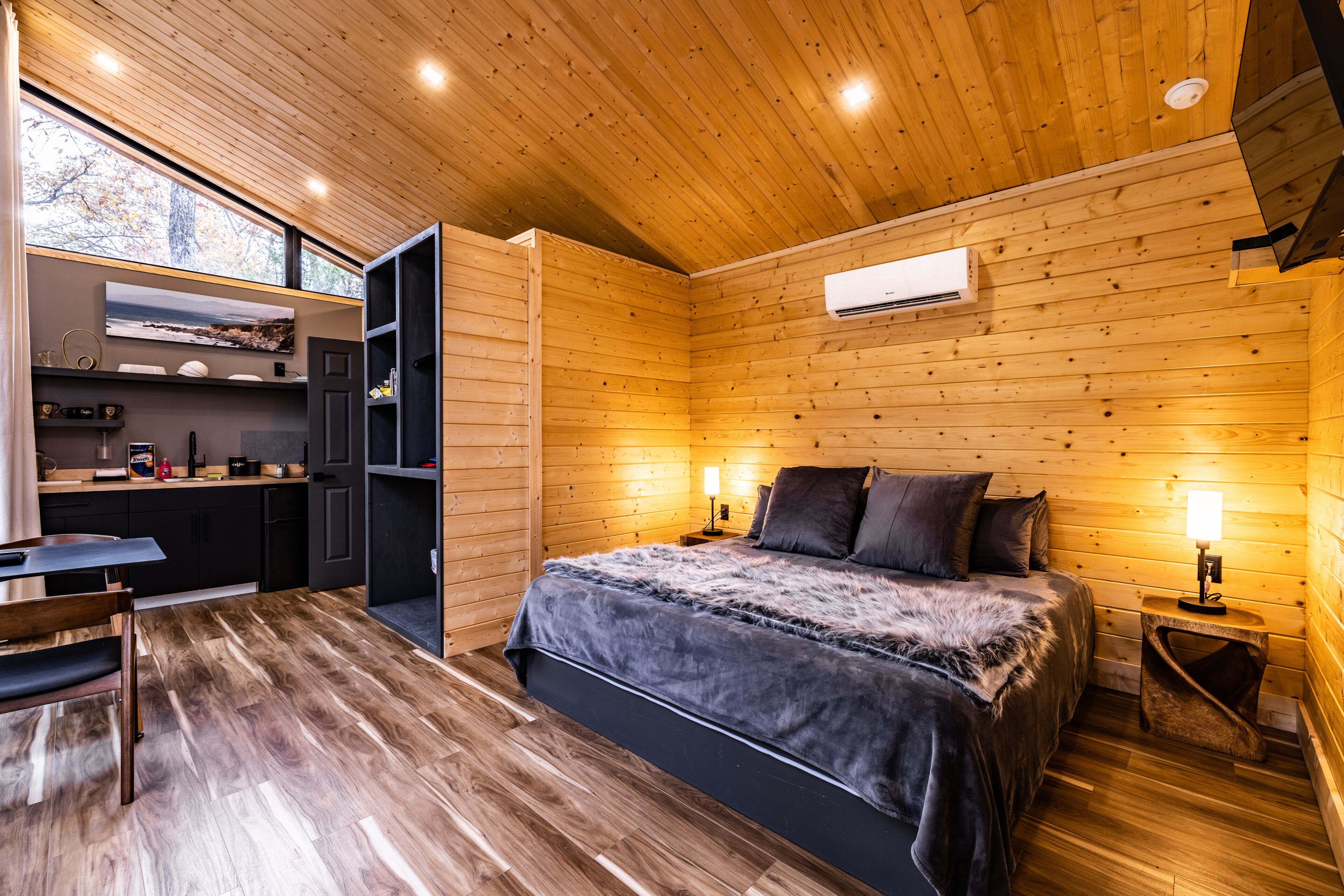 A modern log cabin interior with a wooden ceiling, a bed with a fur throw, a dining area, and a kitchen with a built-in shelf.