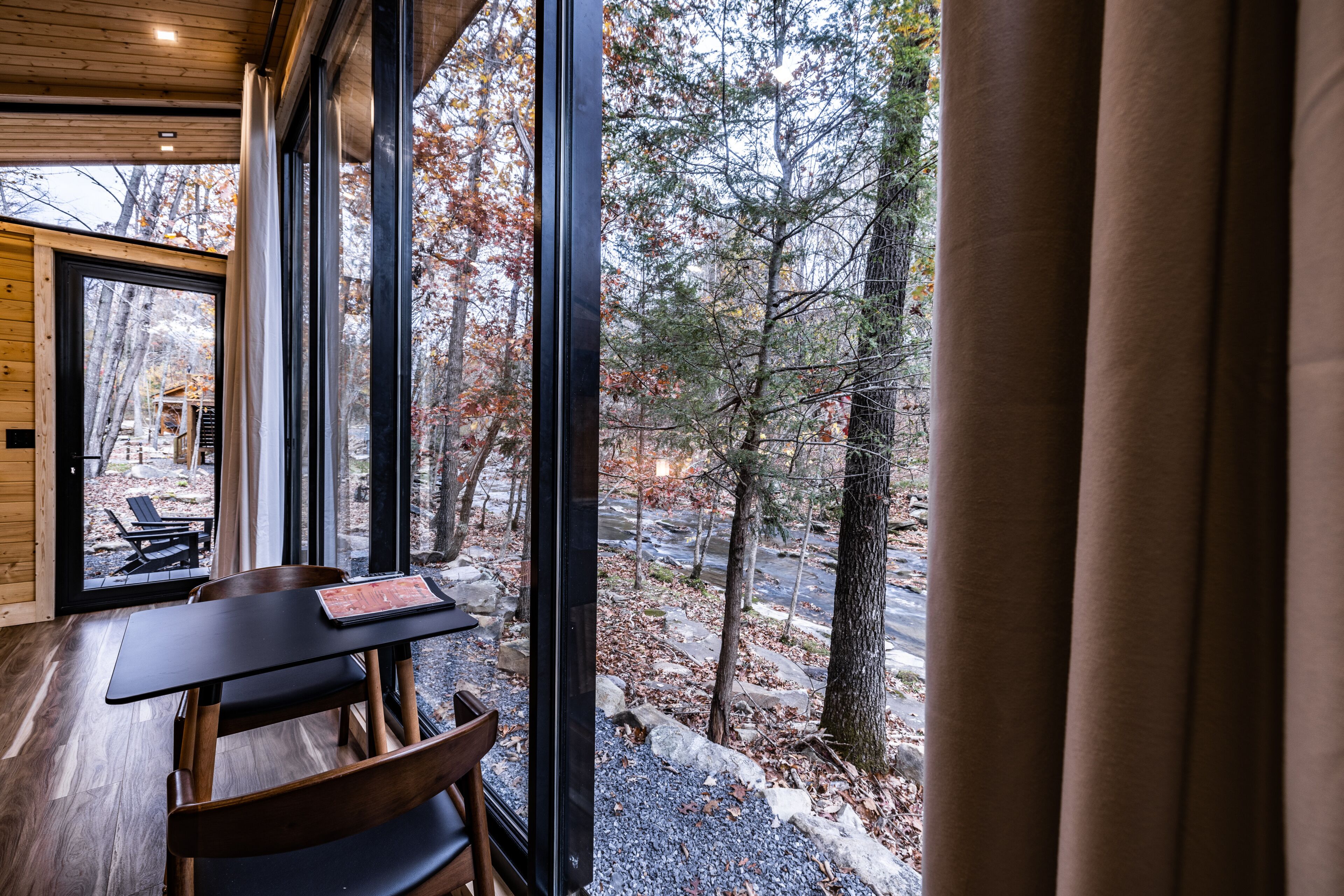 A wooden interior with a table and chairs, large windows, and a view of a forested area.