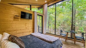 Quarto com cama, televisão, janela com cortinas e vista para árvores.