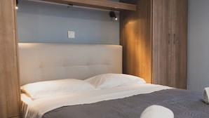 Una cama bien tendida con sábanas blancas y una colcha gris estampada, un armario de madera y un interruptor de luz en la pared.