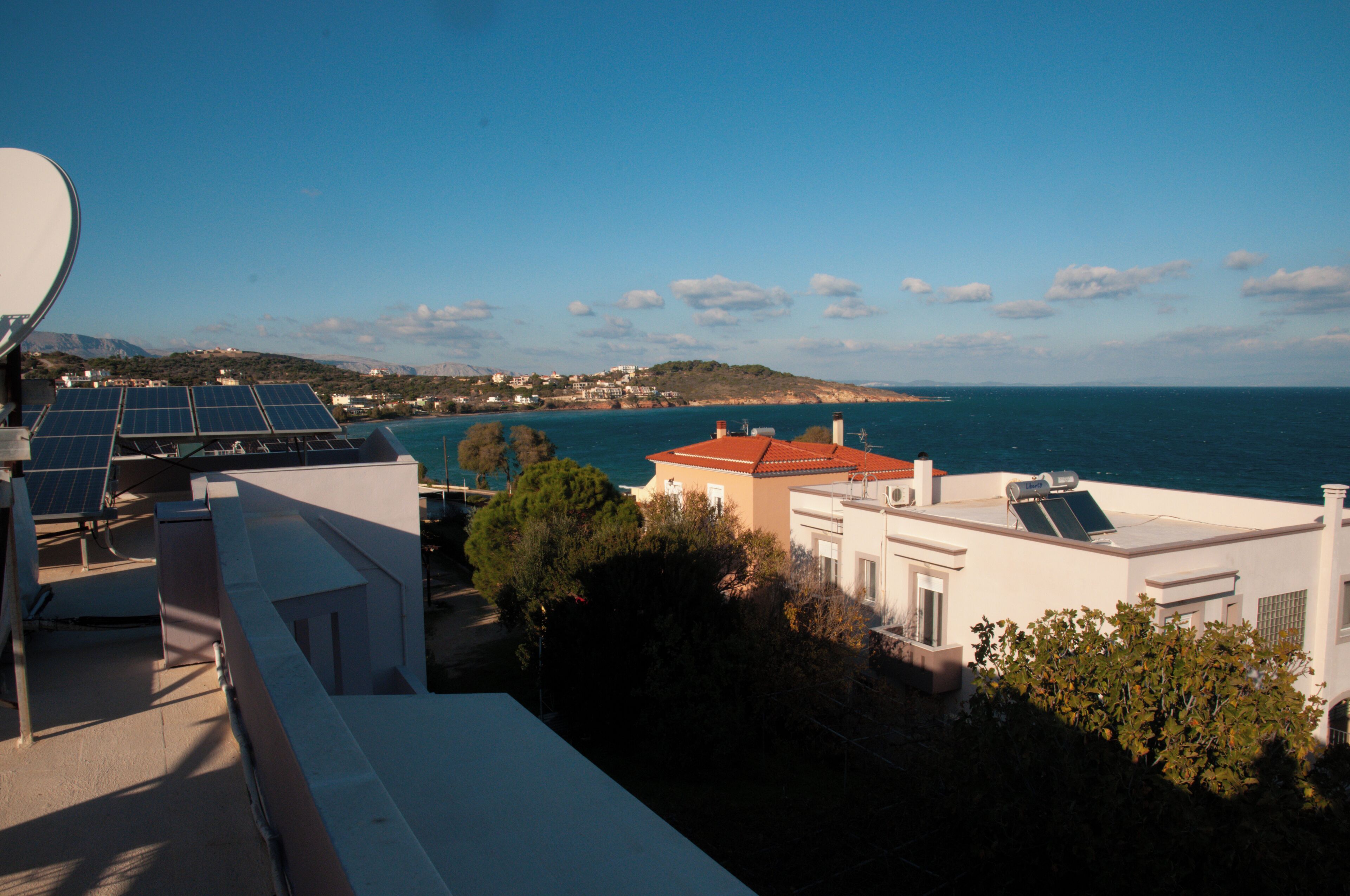 A coastal building with solar panels, a red roof, and a view of the sea and distant hills.
