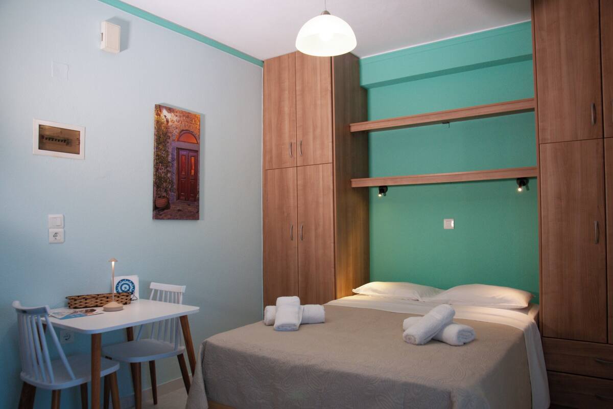 A bedroom with a bed, a desk with chairs, a wall-mounted shelf, and a painting.