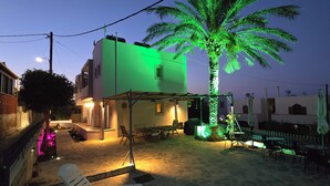 Un patio con una palmera, mobiliario exterior y un edificio con fachada iluminada en verde.