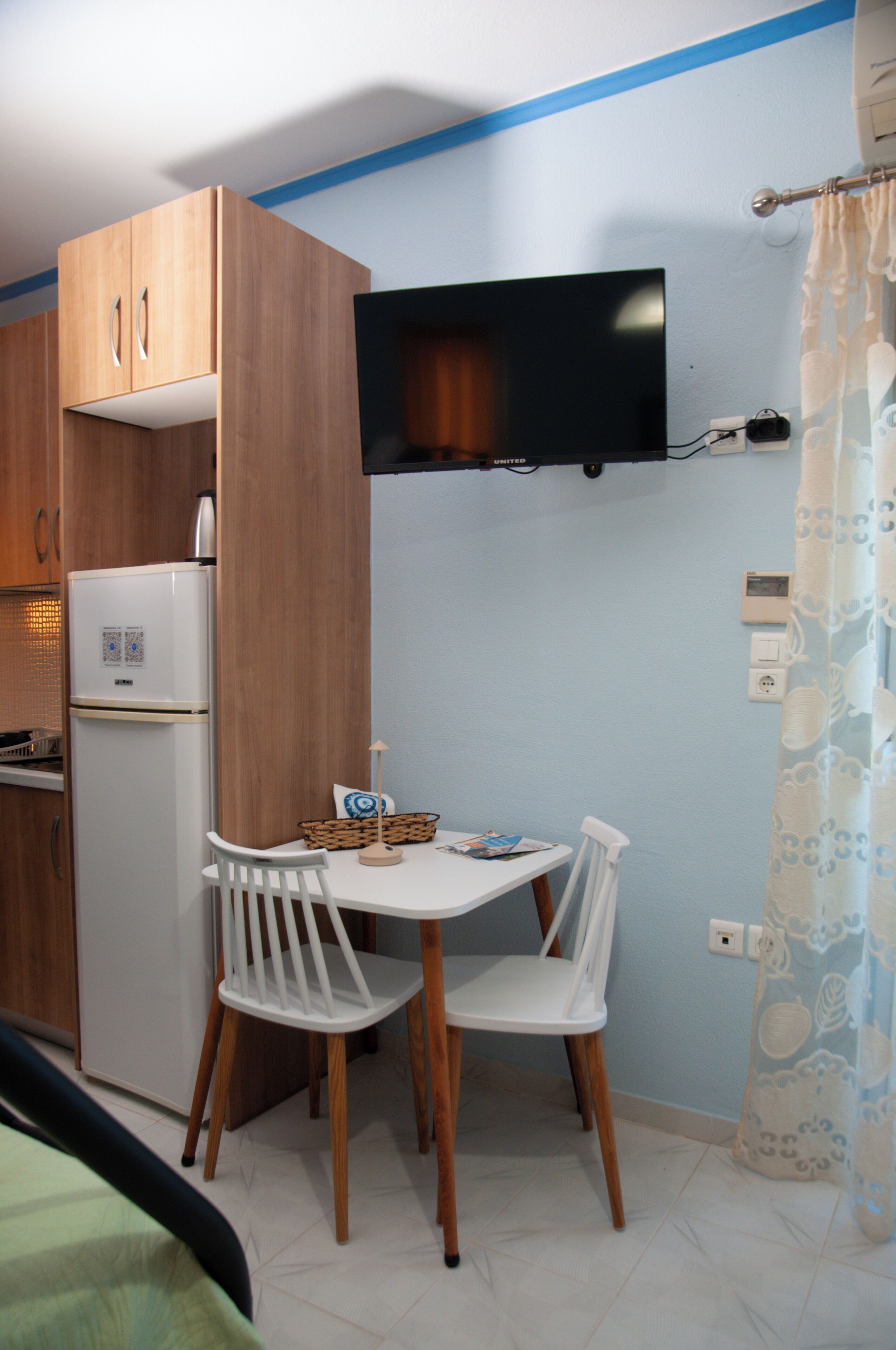 Una cocina compacta con refrigerador, armarios de madera, una mesa de comedor con dos sillas y un televisor montado.