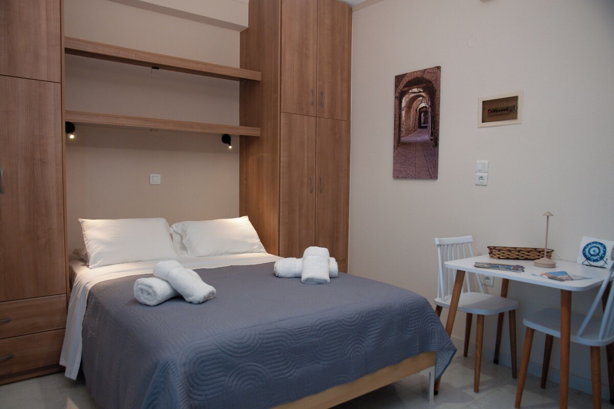 Een netjes opgemaakt bed met witte lakens en opgerolde handdoeken, een houten garderobe, een klein wit tafeltje met stoelen en een aan de muur hangend schilderij.