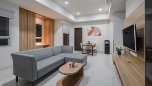 A modern living room with a grey sectional sofa, a wooden coffee table, a TV mounted on the wall, and a dining area with a table and chairs. - Cove Merakey (Cikarang)