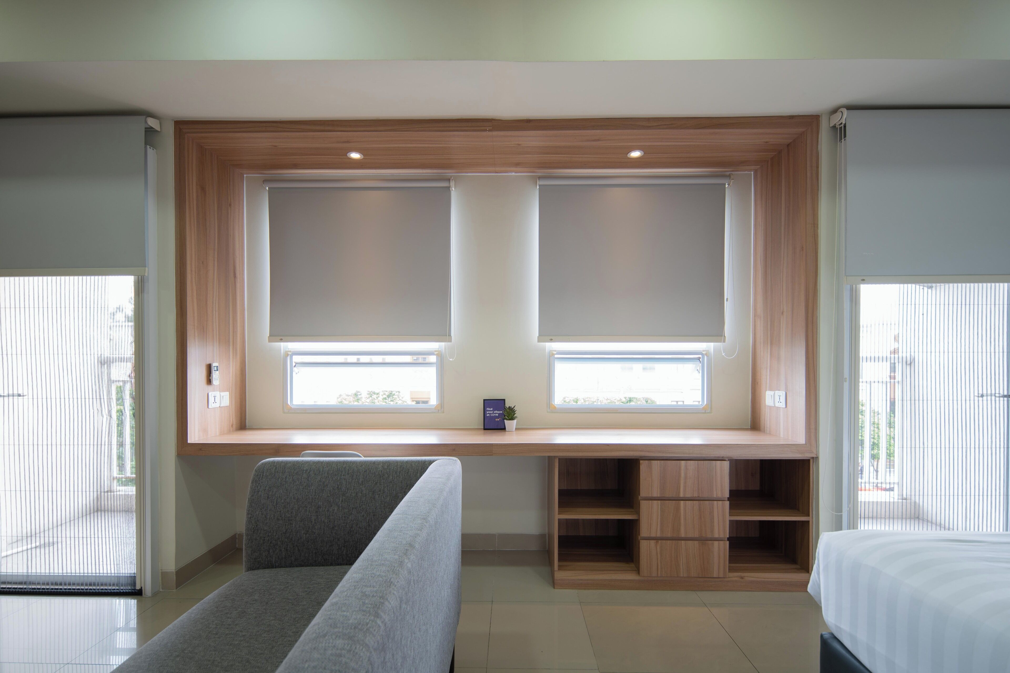 A modern hotel room with a large window, a built-in desk, a grey sofa, and a bed.