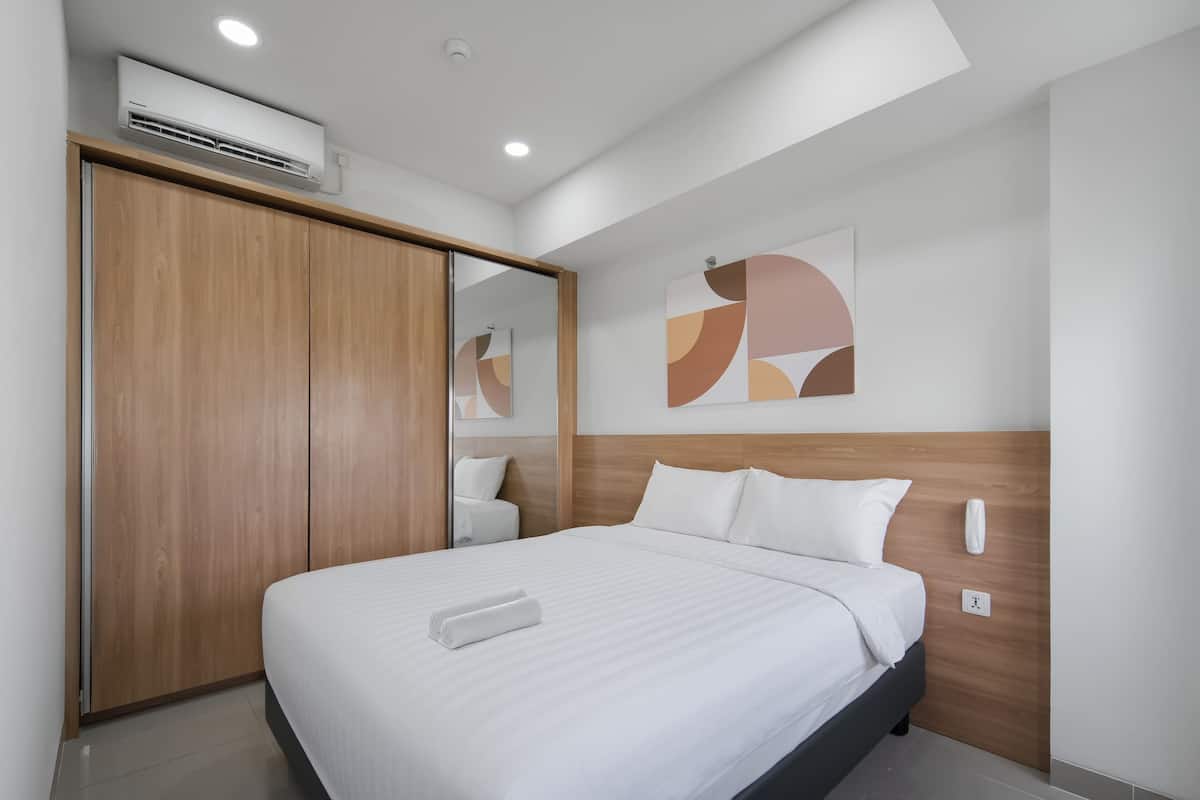 A neatly made bed with a white duvet, a wooden wardrobe, a bedside table with a white lamp, and a wall-mounted phone.
