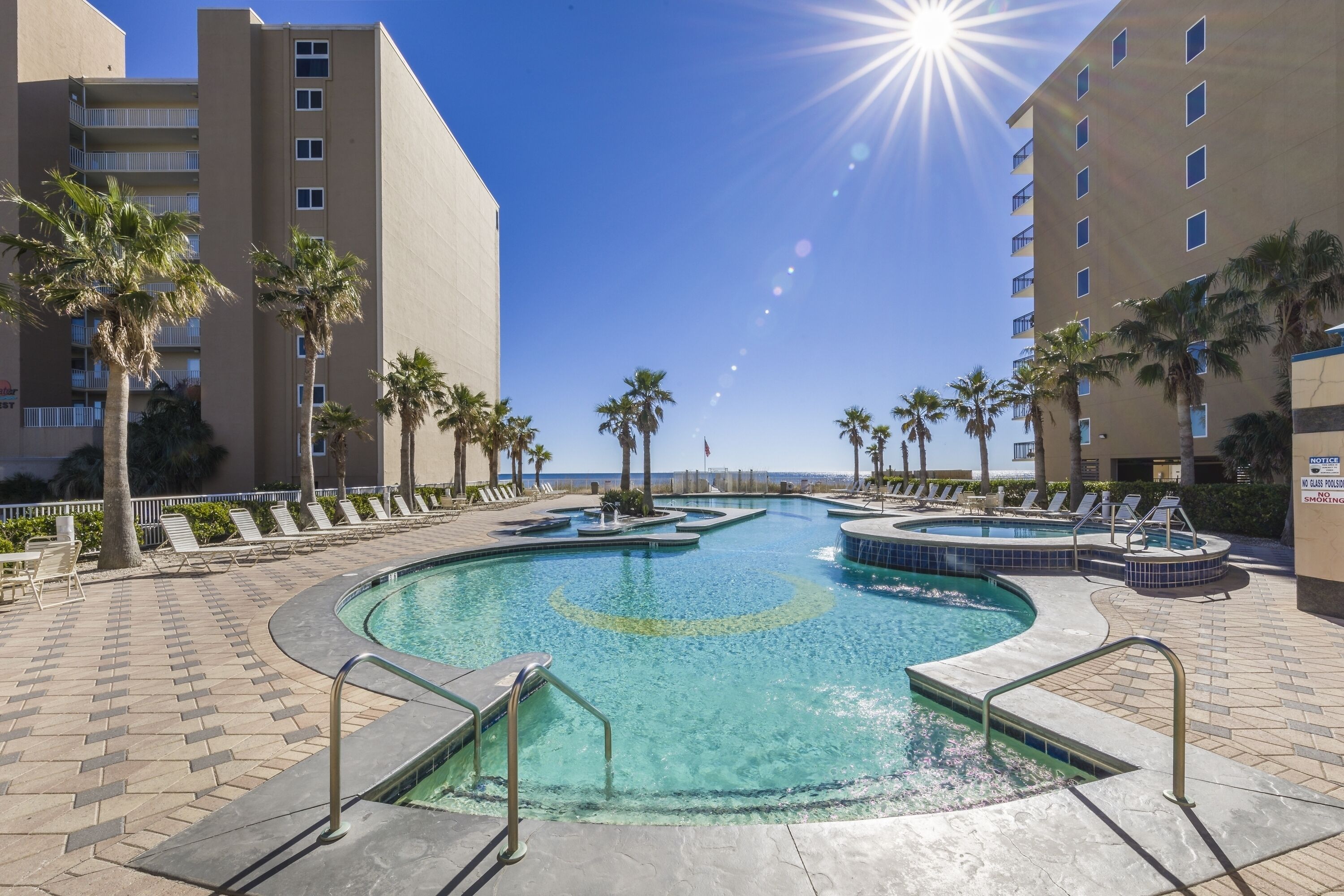 A swimming pool with a sunburst effect, surrounded by palm trees and lounge chairs, with a high-rise building in the background.