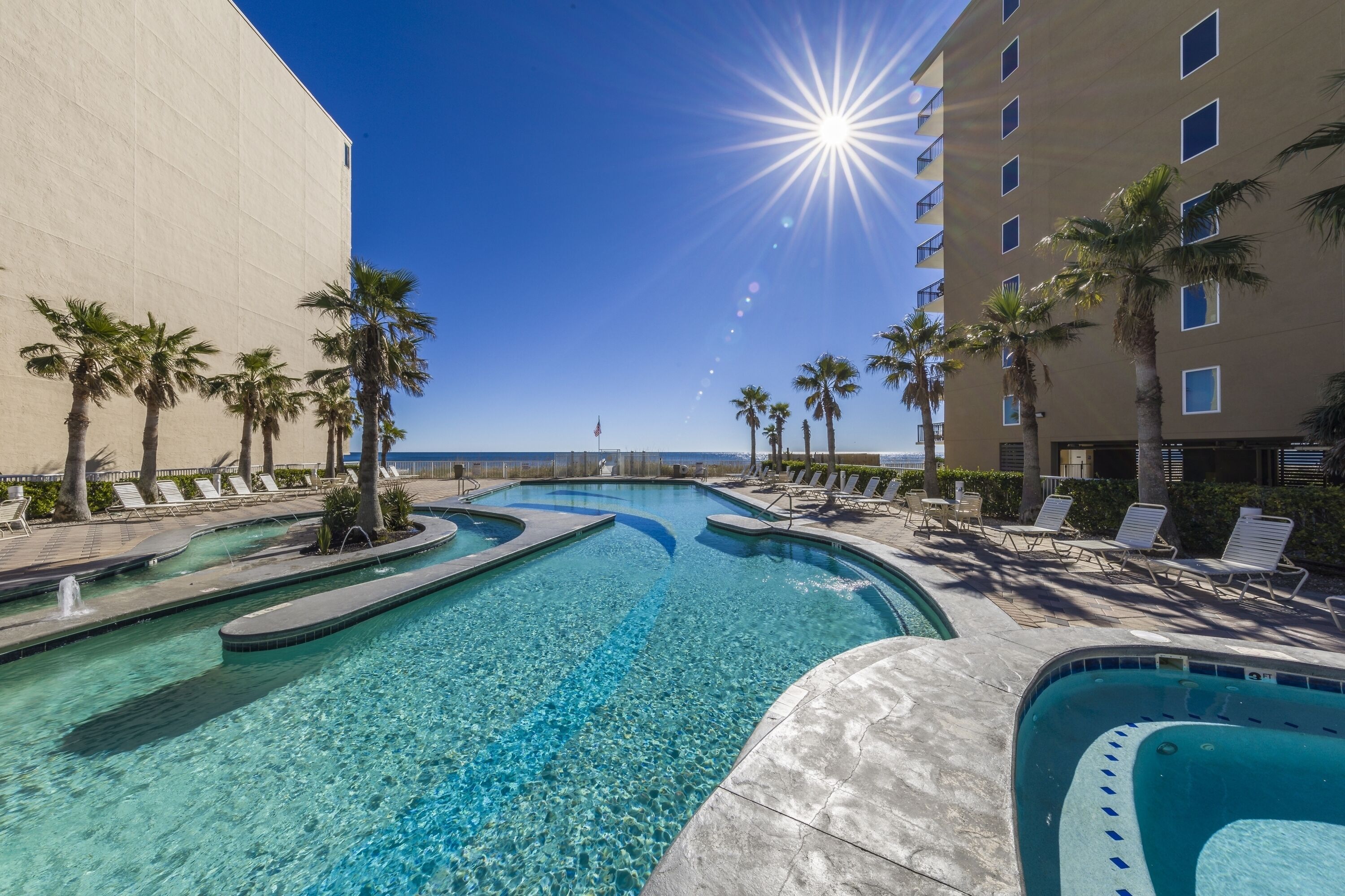 A clear pool with lounge chairs and palm trees, surrounded by a building with balconies.