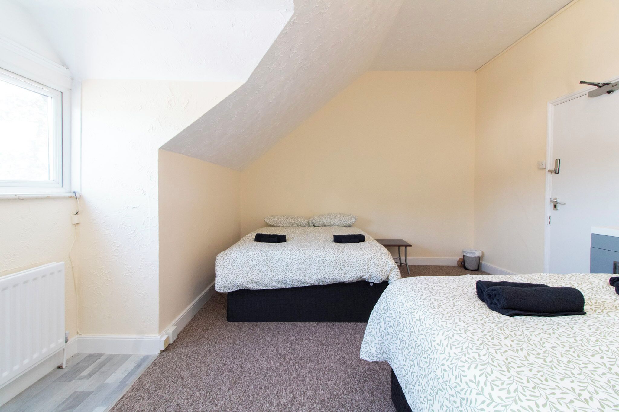 Una habitación pequeña de un dormitorio con techo inclinado, cama con colcha estampada, radiador y ventana.