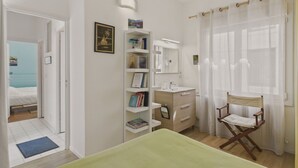 Un dormitorio con cama, mesita de noche, silla, estantería, escritorio y ventana con cortinas.