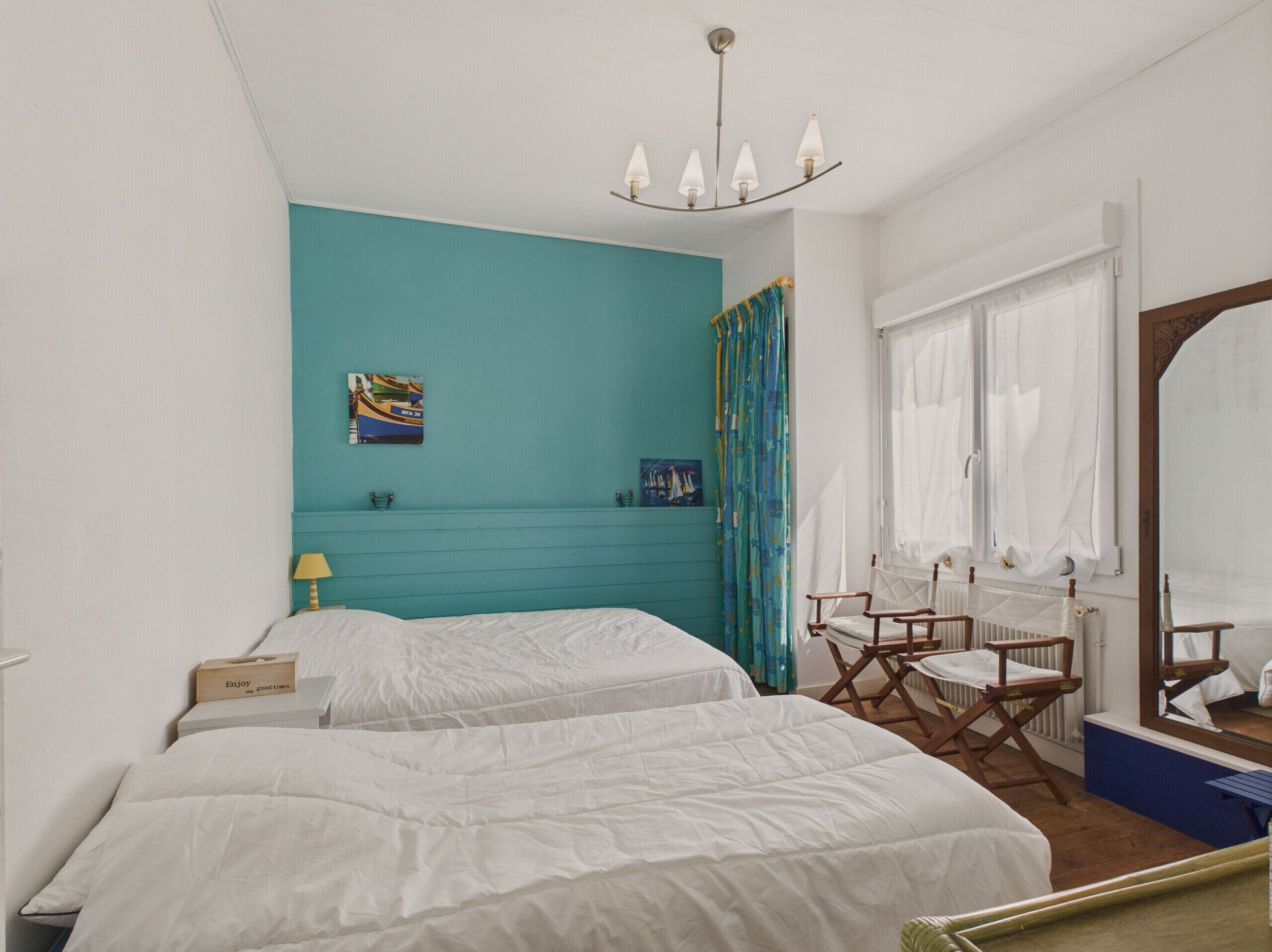 A room with two beds, a window with white curtains, a mirror, and a ceiling light fixture.