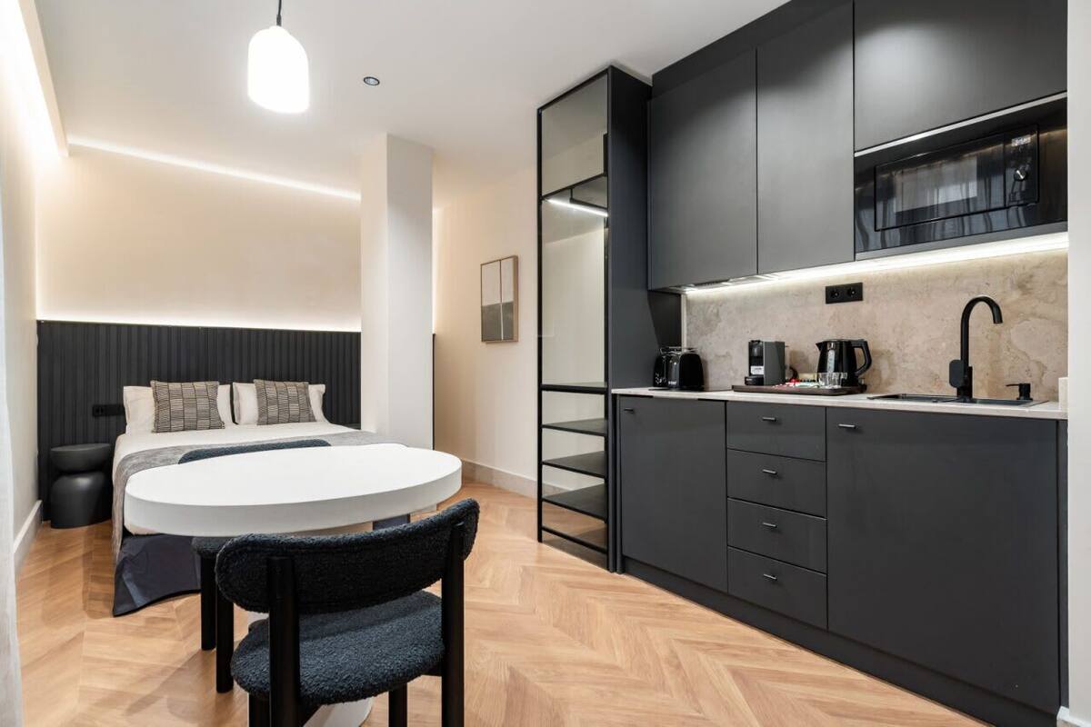 Une chambre d’hôtel moderne équipée d’un lit, d’une table de chevet et d’une petite table ronde avec une chaise. La chambre dispose également d’une kitchenette avec un four à micro-ondes, une machine à café et des placards.