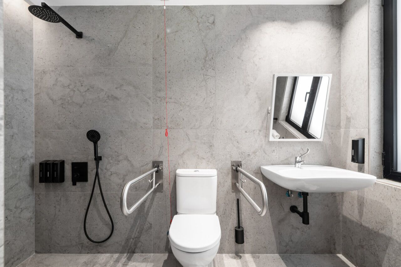 A modern bathroom with a toilet, bidet, and sink, equipped with handrails and a handheld showerhead.