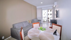 A living room with a grey sofa, a dining table with placemats, a TV mounted on the wall, and a balcony with a view of buildings. - Conforto Localização Imbatível U054 (Rio de Janeiro)