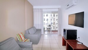 A living room with a grey sofa, a TV on a wooden stand, and a balcony with a view of buildings. - Conforto Localização Imbatível U054 (Rio de Janeiro)