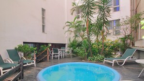 A pool area with lounge chairs and a small table, surrounded by greenery and a building with multiple windows. - Conforto Localização Imbatível U054 (Rio de Janeiro)