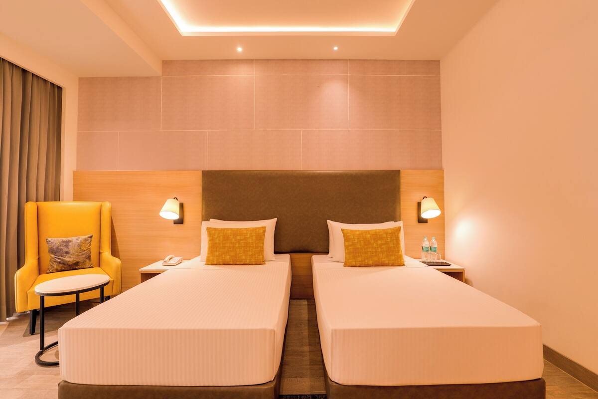 A hotel room with two single beds, a small table, a chair, and a nightstand with a lamp and water bottles.