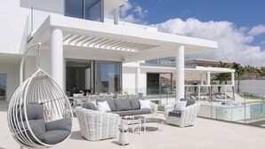 A modern outdoor patio with white wicker furniture, a hanging chair, and a pool area. - Villa Élan (Playa Paraiso)