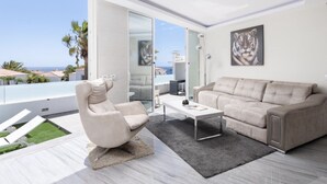 Ein modernes Wohnzimmer mit Sofa, Sessel und Couchtisch, eine Glastür führt zu einem Balkon mit Meerblick.