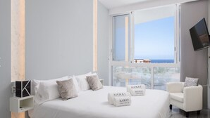 Ein modernes Hotelzimmer mit einem großen Bett, einem weißen Sessel und Meerblick durch ein großes Fenster.