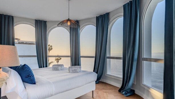 A bedroom with a large bed, a nightstand, a lamp, and a view of the sea through arched windows. - Villa Aurea (El Sauzal)