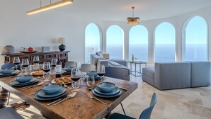 A dining area with a wooden table set for a meal, a sofa, and a view of the ocean through large windows. - Villa Aurea (El Sauzal)