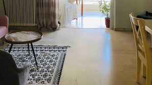 Un salon avec une grande fenêtre, un radiateur, un tapis à motifs, un fauteuil, une table et un luminaire suspendu.