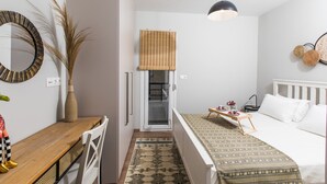 A compact hotel room with a bed, a small desk, a chair, a bedside table with a tray, a hanging light, a window with blinds, and a patterned rug. - Areti Seaview Residence (Rethymno)