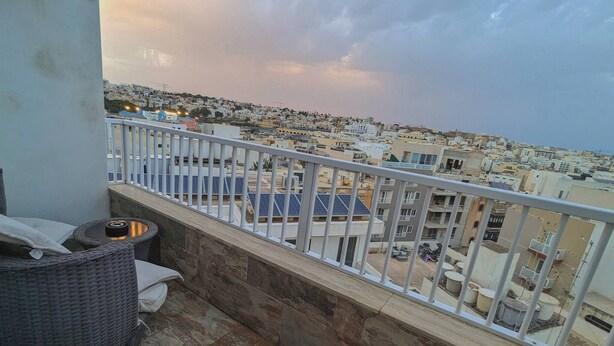A balcony with a view of a densely packed urban area, featuring a stone floor and a wicker chair. - Modern 3 Bedroom Penthouse With Rooftop Views (Msida)