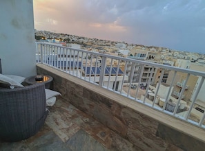 A balcony with a view of a densely packed urban area, featuring a stone floor and a wicker chair. - Modern 3 Bedroom Penthouse With Rooftop Views (Msida)