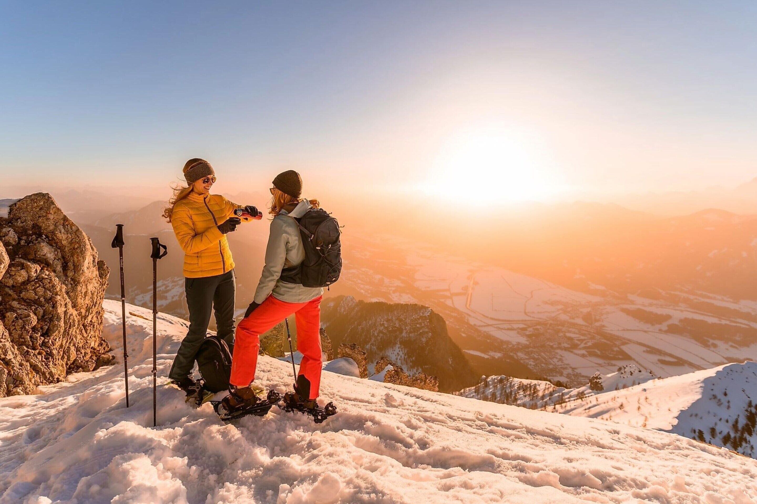 Two people hiking on a snowy mountain with a clear sky and a sunset in the background.