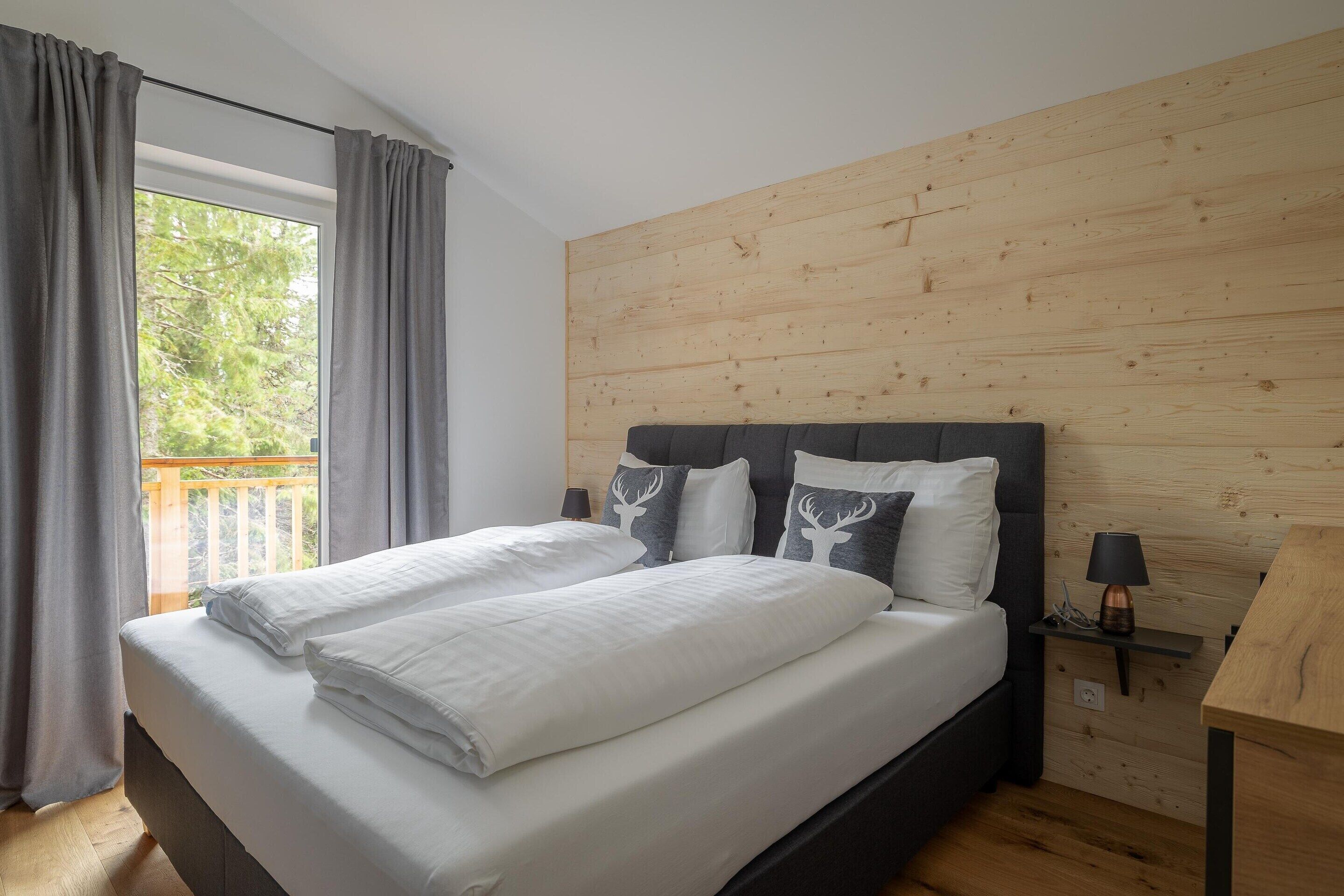 A bedroom with a wooden headboard, a large bed with white linens, a nightstand with a lamp, and a view of greenery through a window with curtains.